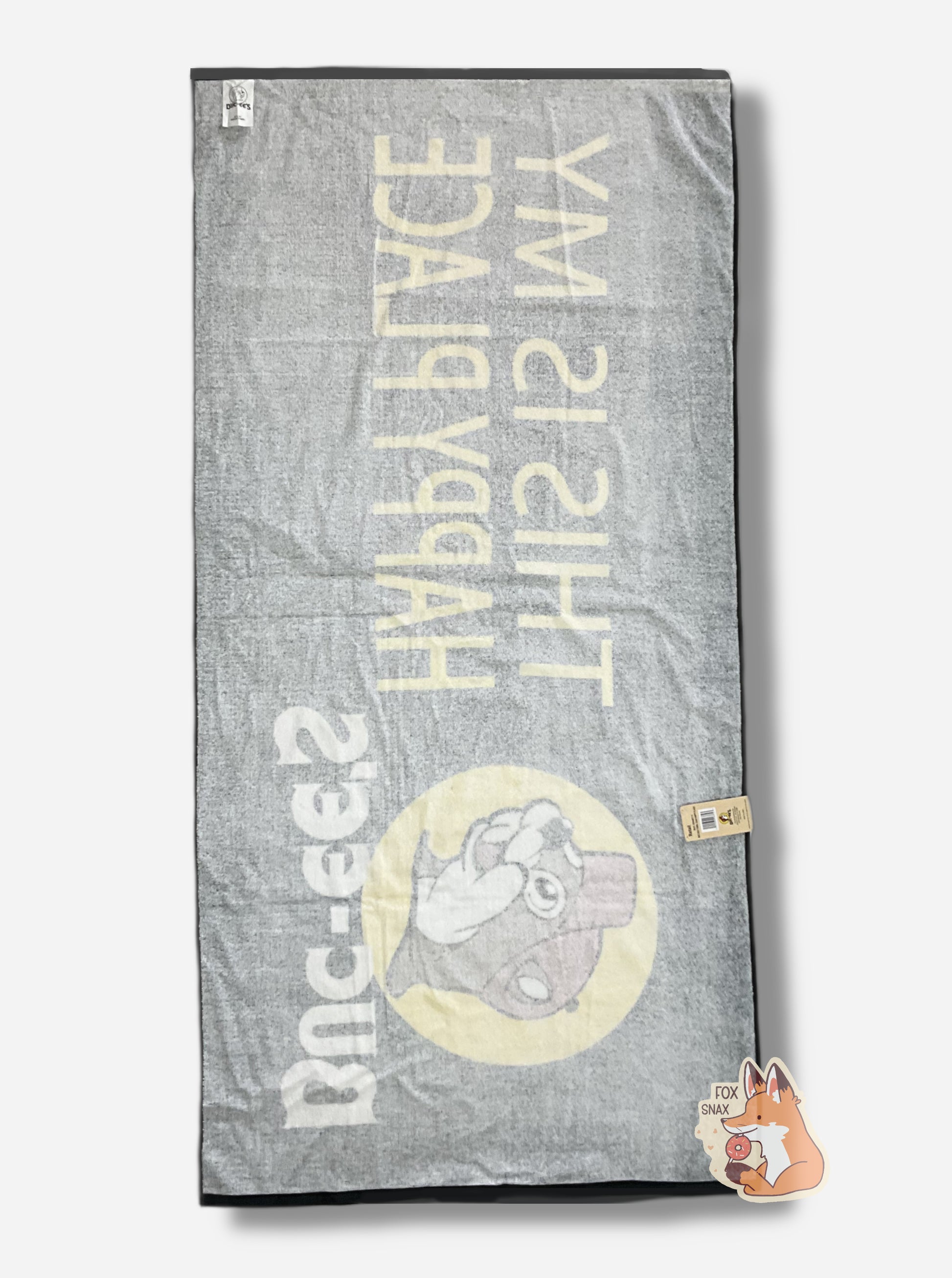 A rotated picture of the back of an  unwrapped, 32x64, fluffy black beach towel.  The back of the towel in white with a black border.  Faintly the front of the towel can be seen; it's (backwards) and adorned with a single Buc-ee The Beaver logo on the middle left.   Buc-ee is framed by a yellow circle, and is wearing a red ball cap. Underneath the logo reads in white text, BUC-EES.  On the right side of the towel, in yellow text, reads THIS IS MY HAPPY PLACE.