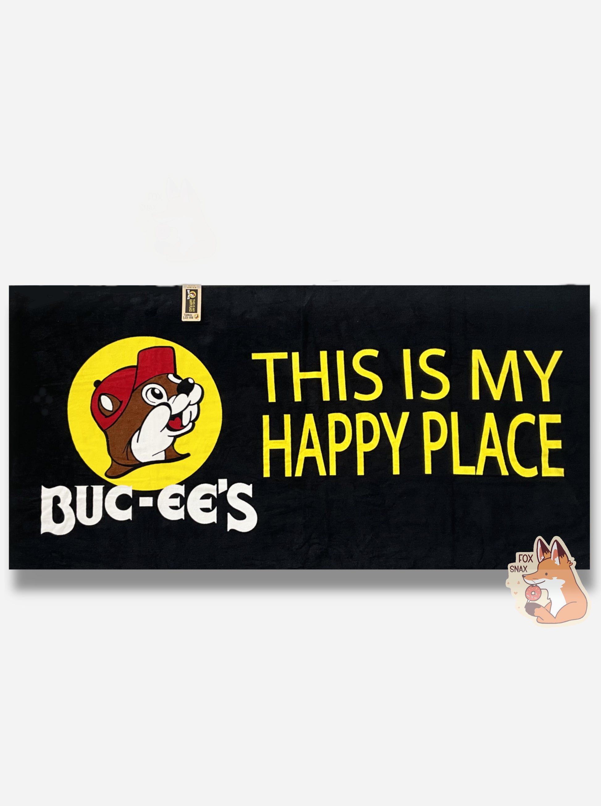 A picture of an unwrapped, 32x64, fluffy black beach towel, adorned with a single Buc-ee The Beaver logo on the middle left.   Buc-ee is framed by a yellow circle, and is wearing a red ball cap. Underneath the logo reads in white text, BUC-EES.  On the right side of the towel, in yellow text, reads THIS IS MY HAPPY PLACE.