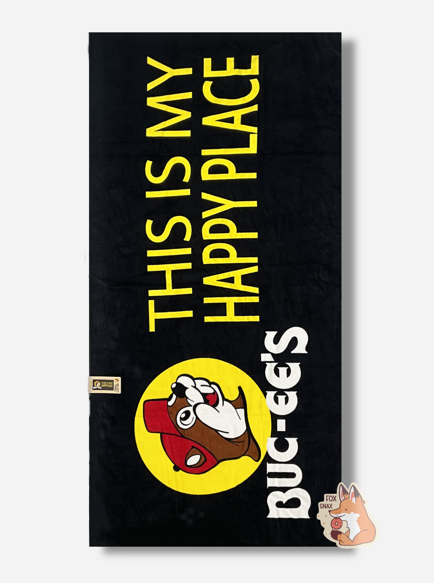 A rotated picture of an unwrapped, 32x64, fluffy black beach towel, adorned with a single Buc-ee The Beaver logo on the middle left.   Buc-ee is framed by a yellow circle, and is wearing a red ball cap. Underneath the logo reads in white text, BUC-EES.  On the right side of the towel, in yellow text, reads THIS IS MY HAPPY PLACE.