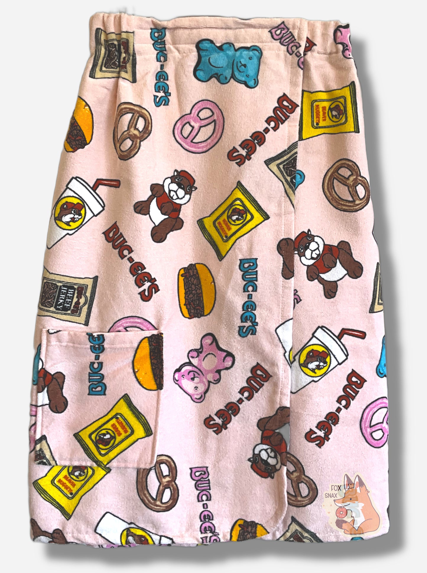 A picture of a pink towel wrap.  The wrap looks fluffy, and has various rotated Buc-ee The Beaver on it, beaver nuggets, a pretzel, the words BUC-EES, a brisket sandwich, a white drink container, and a pink gummy bear icons all over it.  A pocket can be seen on the bottom left of the image, on the wrap.