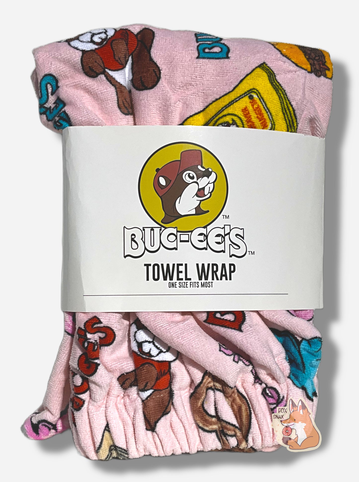 A picture of a wrapped pink towel wrap.  The wrap looks fluffy, and has a picture of Buc-ee The Beaver on it, beaver nuggets, a pretzel, and the words BUC-EES.  A white paper wrap surrounds the middle of the folded wrap, and reads BUC-EE'S TOWEL WRAP.  ONE SIZE FITS MOST underneath the Buc-ee's logo - a cheerful beaver wearing a red ball cap.
