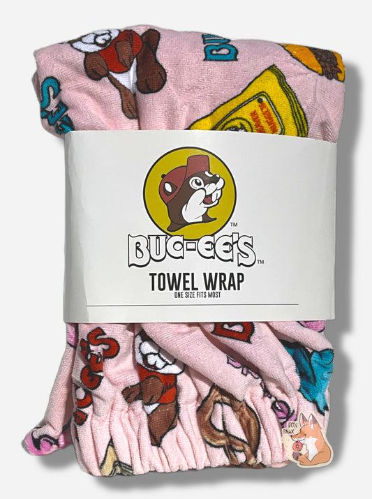 A picture of a wrapped pink towel wrap.  The wrap looks fluffy, and has a picture of Buc-ee The Beaver on it, beaver nuggets, a pretzel, and the words BUC-EES.  A white paper wrap surrounds the middle of the folded wrap, and reads BUC-EE'S TOWEL WRAP.  ONE SIZE FITS MOST underneath the Buc-ee's logo - a cheerful beaver wearing a red ball cap.