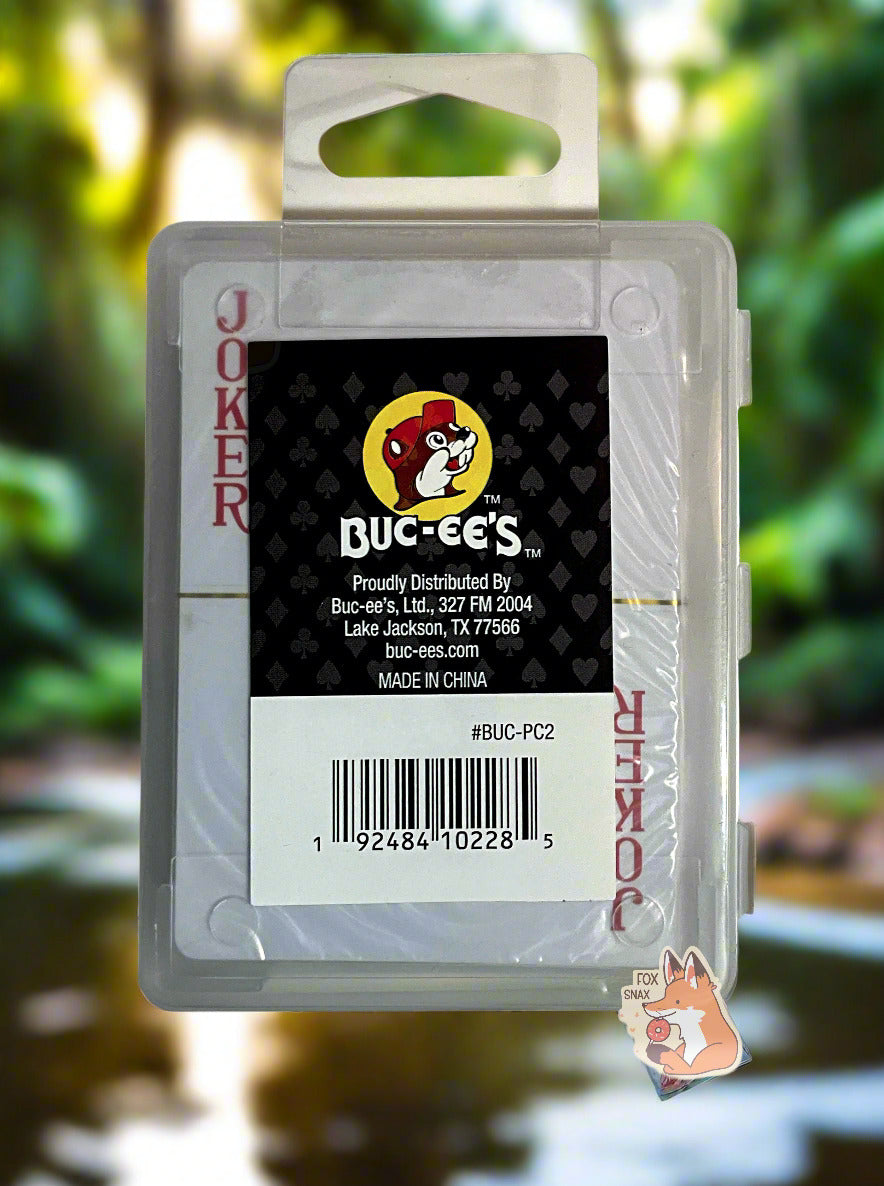 A picture of the back of a pack of Buc-ee's Waterproof Playing Cards.  The pack is plastic, rectangular, and clear.  A joker card can be seen through the plastic.  A small rectangular sticker with the Buc-ee's logo against a hearts/spades/diamonds/clubs background can be seen.  The cards are suspended in midair, and the background is of a forest with a river nearby.