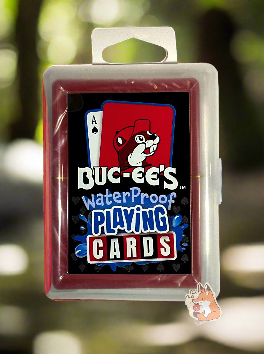 A picture of a pack of Buc-ee's Waterproof Playing Cards.  The pack is plastic, rectangular, and clear.  The front of the pack has two cards at the top - the first the ace of spades, the second a picture of Buc-ee the Beaver against a red background.  Underneath the cards reads: BUC-EE'S WATERPROOF PLAYING CARDS, each word in a different font.  The words PLAYING CARDS are surrounded by splashing blue water.  The cards are suspended in midair with a forest background.