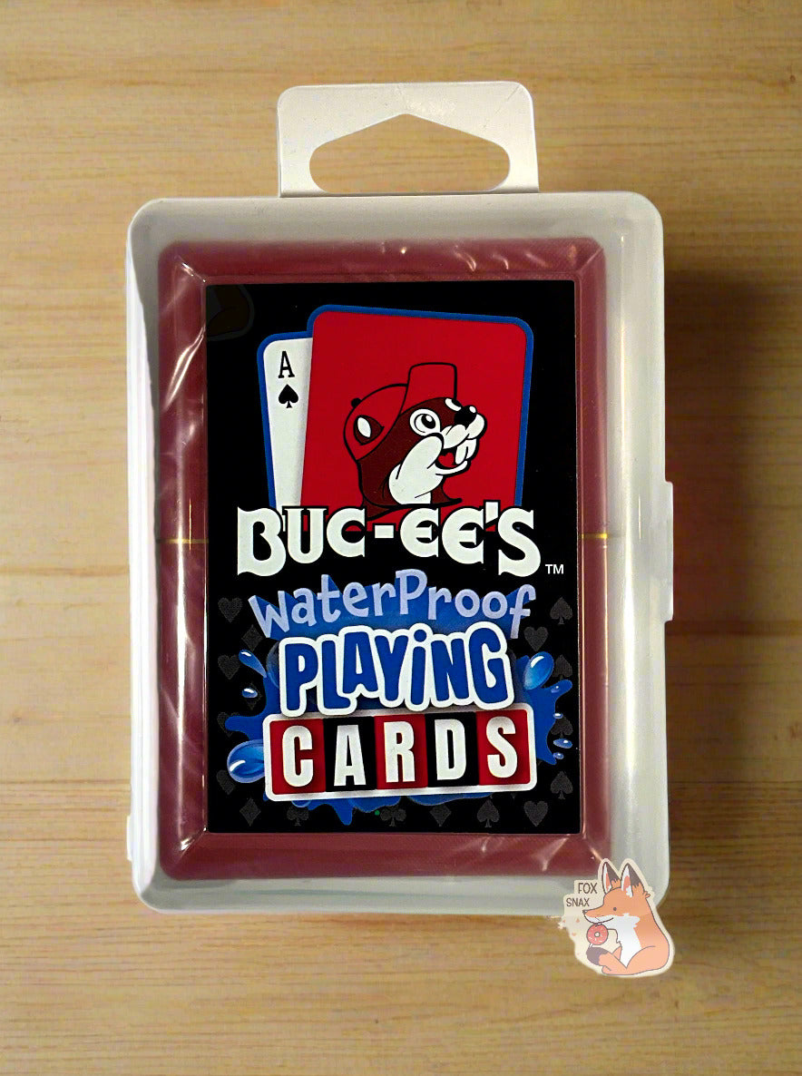 A picture of a pack of Buc-ee's Waterproof Playing Cards.  The pack is plastic, rectangular, and clear.  The front of the pack has two cards at the top - the first the ace of spades, the second a picture of Buc-ee the Beaver against a red background.  Underneath the cards reads: BUC-EE'S WATERPROOF PLAYING CARDS, each word in a different font.  The words PLAYING CARDS are surrounded by splashing blue water.  The cards are sitting on a light wooden table.