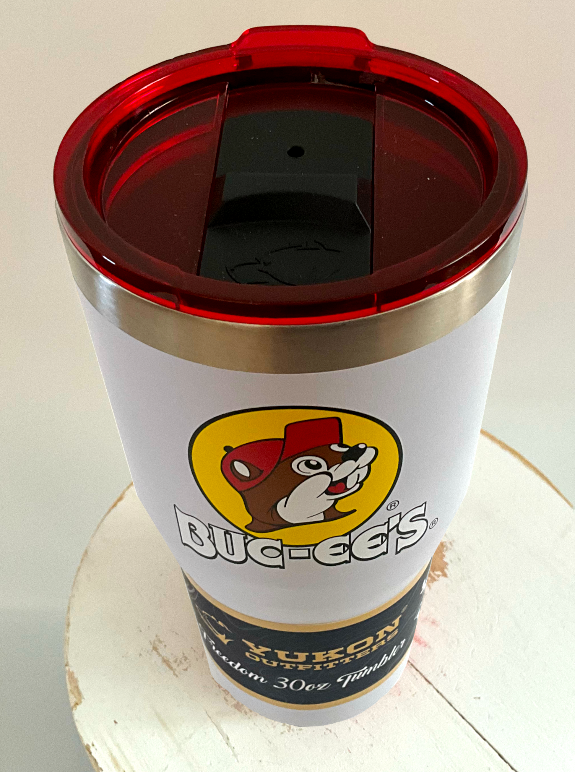 A 3/4 view of a picture of a tall, white, tumbler, with a big picture of Buc-ee The Beaver on it.  The surface is smooth and white, as it is white paint covering stainless steel.  There is no handle.  The top is red plastic, and shows a black slider in the middle with the imprint of the Yukon bear's head on it.  The top of the mug is lined with a stainless steel band.  A paper wrapper around the bottom reads: Yukon Outfitters.  Freedom 30 oz Tumbler.  The tumbler is sitting on a white circular stool. 